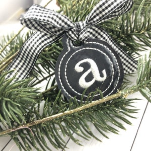 May include: A black and white checked ribbon tied around a black felt ornament with a white embroidered letter "A" on it.