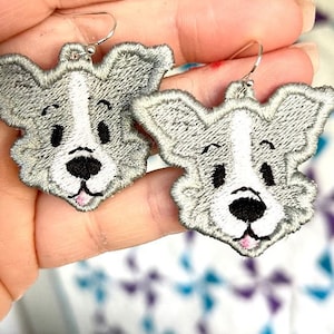 Może przedstawiać: Para srebrnych kolczyków z haftowanym wzorem psa w kolorze szarym i białym. Pies ma białą twarz z czarnymi oczami i czarnym nosem.