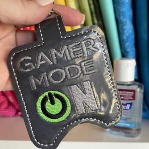 以下が含まれることがあります： グレーの布製のキーホルダーで、銀色の刺繍で「GAMER MODE ON」と書いてあります。テキストの下には緑色の電源ボタンのシンボルが刺繍されています。