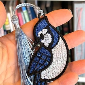 May include: Embroidered blue jay bookmark with a light blue tassel. The bird features blue, white, and black stitching. The bookmark is held in a hand, showcasing the intricate details of the design.