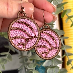 May include: A pair of pink and brown dangle earrings with a concha design. The earrings are made of fabric and have a brown wire loop.