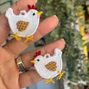 May include: A pair of white chicken-shaped earrings with red combs, yellow beaks, and yellow feet. The earrings are made of embroidered fabric and have gold-coloured hoops.