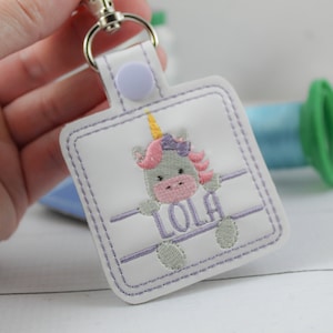 May include: White key chain with a purple unicorn with a pink nose and a yellow horn. The unicorn is holding a banner with the name "Lola" embroidered on it.