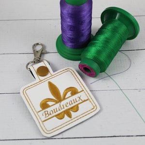 May include: White square keychain with a gold embroidered fleur-de-lis and the name "Boudreaux" underneath. The keychain is attached to a silver key ring. Two spools of thread, one purple and one green, are in the background.
