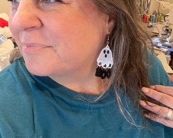 May include: A pair of dangle earrings featuring a white ghost with a black face and the word "BOO" in black letters.