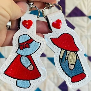 May include: Two keychains featuring embroidered designs. One depicts a figure in a red dress and blue hat, the other a figure in a red hat and blue overalls. Each keychain has a red heart and a silver clasp.