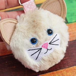 May include: A white fluffy cat keychain with blue eyes and a pink nose. The cat has embroidered whiskers and a white mouth. The keychain has a pink and white polka dot ribbon attached to it.
