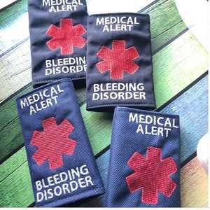 May include: Four black and blue fabric medical alert bracelets with a red star of life and the text "Medical Alert" and "Bleeding Disorder".