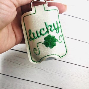 Peut inclure: Porte-gel hydroalcoolique blanc avec un trèfle vert et le mot "lucky" brodé dessus. Le porte-gel est attaché à un porte-clés argenté.