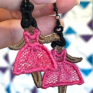 May include: A pair of dangle earrings featuring two embroidered figures of people wearing pink dresses. The figures have brown skin and black hair. The earrings are made with black metal hooks.