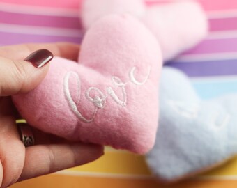 Könnte beinhalten: Ein rosa, herzförmiges Plüschtier mit dem Wort "Love" in weiß aufgestickt. Das Spielzeug wird von einer Hand gehalten.
