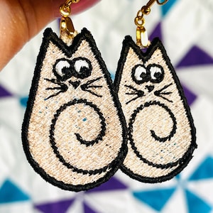 Puede incluir: Un par de pendientes con forma de gato con la cola enroscada, ojos negros y un diseño bordado en blanco y beige.