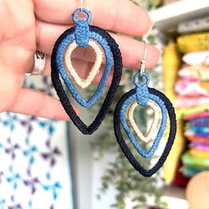 Peut inclure: Une paire de boucles d'oreilles pendantes à trois couches de tissu. La couche extérieure est noire, la couche intermédiaire est bleue et la couche intérieure est crème. Les boucles d'oreilles ont la forme de larmes.