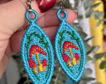 Op de afbeelding: Een paar turquoise blauwe oorhangers met geborduurde rode en gele paddenstoelen op een groen rankontwerp.