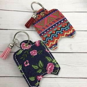 May include: Two hand sanitizer holders with keychains and tassels. One holder is red with a geometric pattern and the other is blue with a floral pattern.