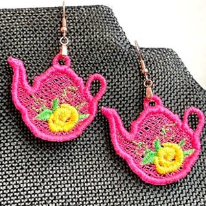 以下が含まれることがあります： 黄色い花の刺繍が施されたピンクのティーポット型のイヤリングのペア。イヤリングは繊細なレースのような素材で作られており、ヴィンテージな魅力があります。