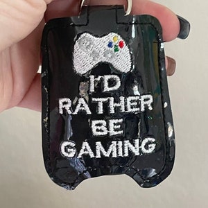 Peut inclure: Porte-gel hydroalcoolique noir avec une manette de jeu vidéo brodée en blanc et le texte "I'd Rather Be Gaming".