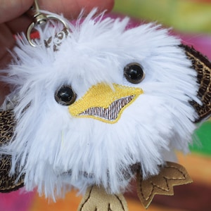 May include: White fluffy plush eagle keychain with brown leather wings and a yellow beak. The eagle has black eyes and a friendly expression.