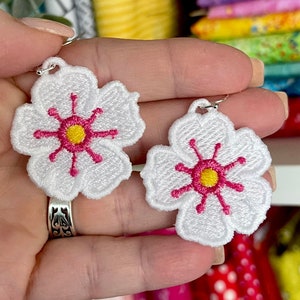 May include: A pair of white flower-shaped earrings with embroidered details. Each earring features a white flower with a yellow center and pink accents. The earrings are held up by silver hooks.