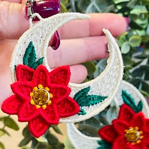May include: Embroidered crescent moon earrings with a red flower and green leaves. The earrings have a cream-colored base with a red flower and a yellow gem in the center. The earrings are a unique accessory.