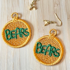 May include: A pair of gold-toned earrings with a round, gold fabric base and green embroidered text that reads "BEARS".