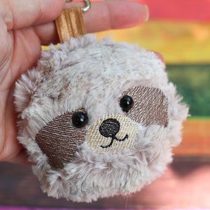 Puede incluir: Un llavero de peluche marrón y blanco con forma de perezoso con ojos bordados y una boca sonriente. El llavero se sostiene en una mano sobre un fondo de arcoíris.