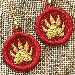 May include: Embroidered red earrings with a gold paw print design. The earrings have a circular shape with a gold-colored hook. The paw print is in the center of the red circle. The earrings are displayed on a textured, neutral-colored surface.