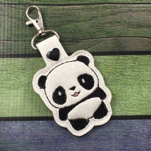 May include: White keychain with a black and white panda bear design. The panda is smiling and has a black heart on the top of the keychain.