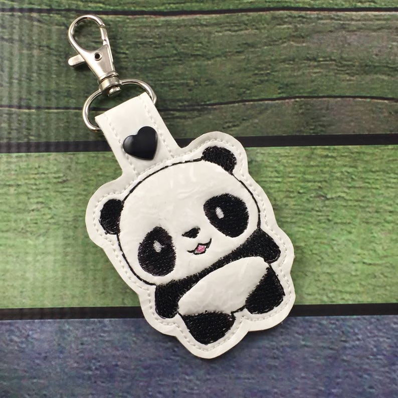 May include: White keychain with a black and white panda bear design. The panda is smiling and has a black heart on the top of the keychain.
