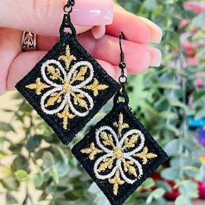 May include: A pair of black square earrings with gold and white embroidery. The earrings feature a floral design.