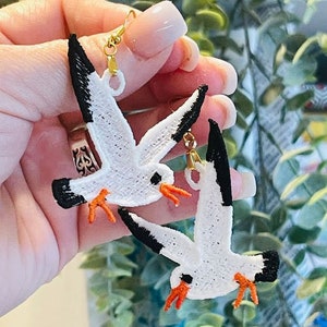 May include: A pair of white seagull earrings with black accents and orange beaks. The earrings are made of fabric and have gold-colored hooks.