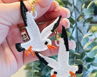 May include: A pair of white seagull earrings with black accents and orange beaks. The earrings are made of fabric and have gold-colored hooks.