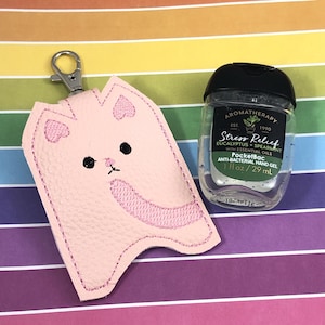 Peut inclure: Porte-clés en cuir rose en forme de chat avec un anneau de clé argenté. Le porte-clés est conçu pour contenir une petite bouteille de désinfectant pour les mains. La bouteille est transparente et a un bouchon noir. L'étiquette de la bouteille indique "Stress Relief Eucalyptus + Spearmint with Essential Oils PocketBac Anti-Bacterial Hand Gel 1 fl oz / 29 ml"