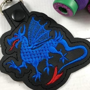 May include: A black vinyl keychain featuring an embroidered blue dragon with a red tongue and tail tip. The dragon is stitched onto a black base with a scalloped edge, secured by a black snap button and a silver metal ring at the top.