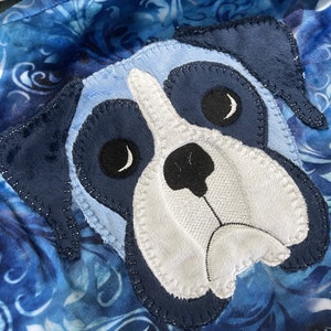 Puede incluir: Una aplicación de perro de peluche azul y blanco con ojos negros y nariz negra. La aplicación está cosida sobre una tela azul y blanca con un patrón floral.