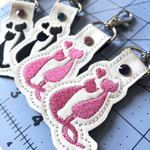 Puede incluir: Cuatro llaveros de cuero blanco con siluetas de gatos bordadas en rosa y negro. Cada llavero tiene un adorno de strass.