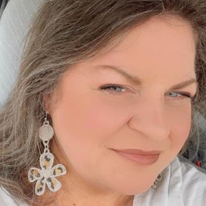 May include: A woman wearing a white shirt with a silver chain necklace and a pair of dangle earrings. The earrings are silver with a floral design and a leopard print pattern.