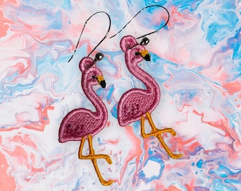 May include: A pair of flamingo-shaped earrings. The earrings are embroidered with a deep pink thread, featuring black beaks and yellow legs. The background has a marbled effect with pink, blue, and white colors.