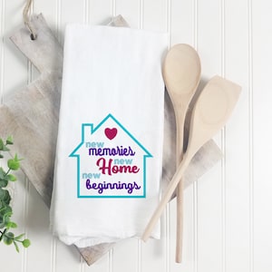 May include: White kitchen towel with a turquoise house design and the text "new memories new home new beginnings" in purple and pink lettering. A red heart is inside the house.