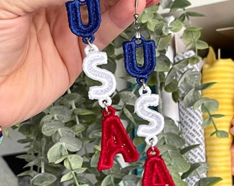 Può includere: Un paio di orecchini pendenti con le lettere "USA" ricamate in blu, bianco e rosso. Gli orecchini sono in argento e hanno una chiusura a gancio.