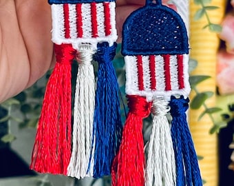 May include: A pair of red, white, and blue tassel earrings with an American flag design. The earrings are made of fabric and have a silver hook closure.