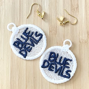 May include: A pair of white embroidered earrings with the text "BLUE DEVILS" in navy blue thread. The earrings are attached to gold hooks.