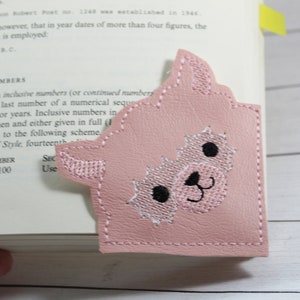 May include: A pink llama bookmark with black eyes and a white nose. The bookmark is made of felt and has a stitched edge.