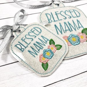 May include: Two white glitter gift tags with the text "Blessed Mama" and blue and pink floral embroidery.