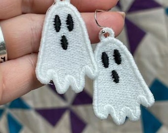 May include: A pair of white ghost-shaped earrings with black eyes and a smiling mouth. The earrings are made of fabric and are hanging from silver hooks.