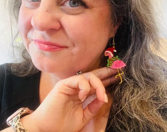 May include: A close-up of a person wearing a pair of flamingo earrings with a Christmas theme. The earrings feature pink flamingos with green accents and Santa hats. The person is also wearing a leopard print watch band.