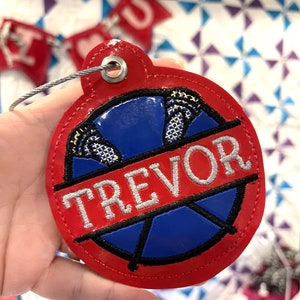 May include: Red round fabric keychain with a blue and black basketball design and the name "TREVOR" in white embroidery.