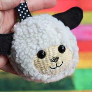 May include: A white fluffy sheep keychain with black ears and eyes. The keychain has a black and white polka dot ribbon attached.