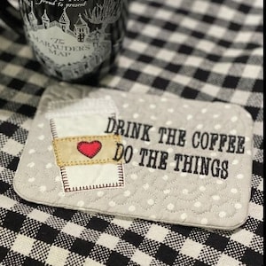 May include: A gray and white quilted coaster with the text "Drink the coffee do the things" and a stylized image of a coffee cup with a red heart.