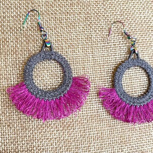 May include: A pair of gray crocheted hoop earrings with a pink fringe. The earrings have a simple, elegant design.